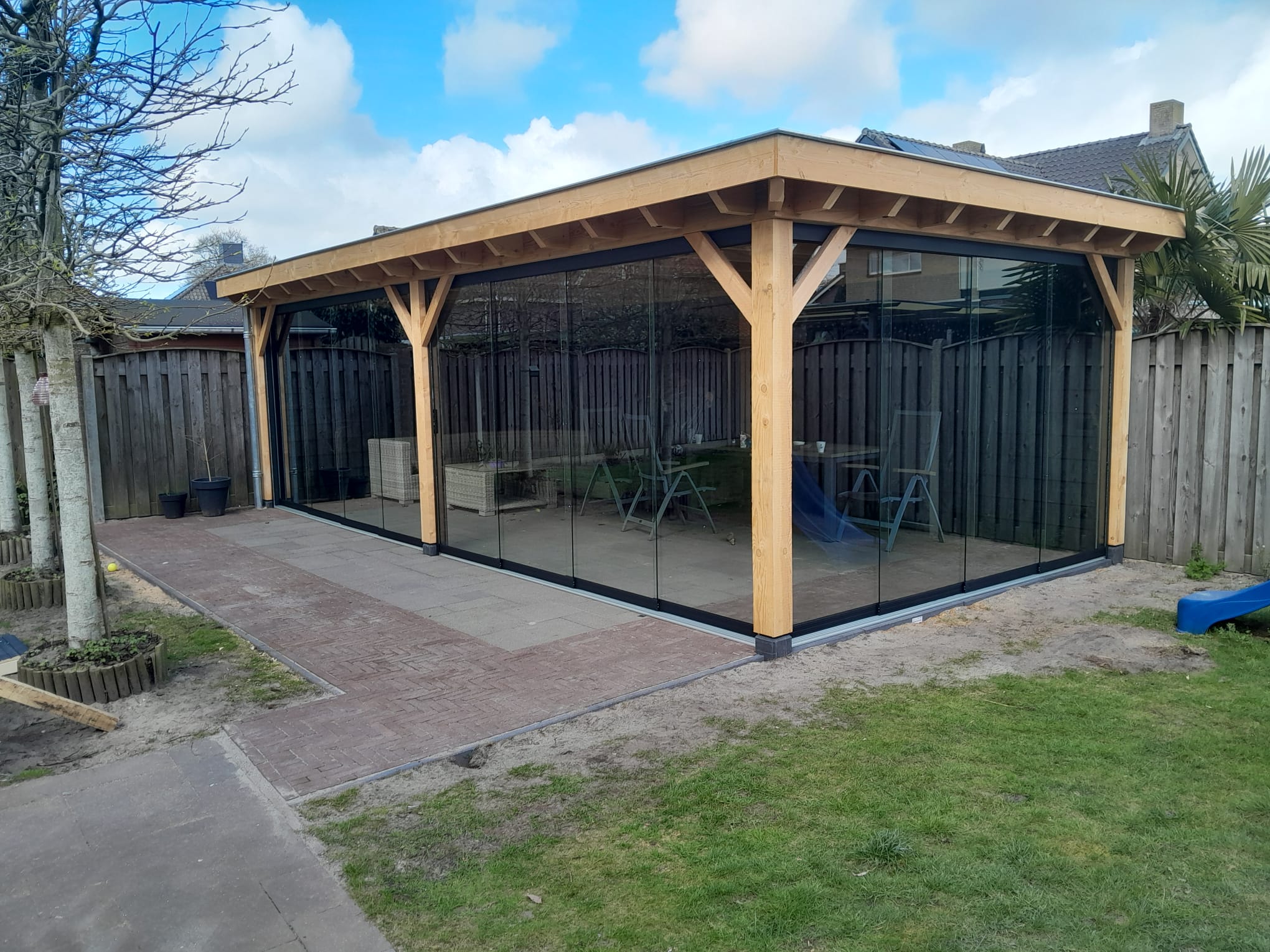 Glazen schuifwand onder houten overkapping met zwarte profielen in tuin